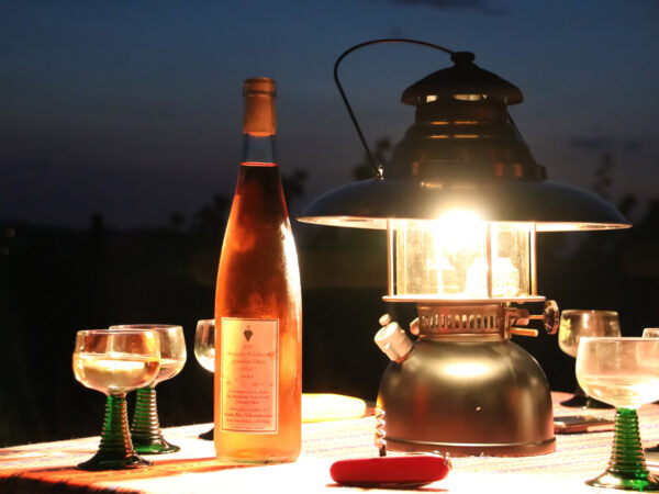 Die Sonne ist längst untergegangen. Auf dem Tisch stehen Weingläser, eine Flasche Rosé-Wein sowie eine Petromax-Petroleumlampe. Ein Korkenzieher liegt bereit. Das Licht der Petroleumlampe verleiht dem angerichteten Weingedeck eine romantische Atmosphäre.