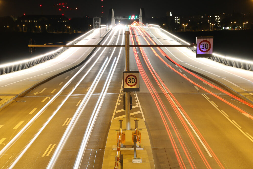 Oberhalb des Ausgangs des Waldschlösschentunnels stehend führt der Blick fahrbahnbegleitend über die Dresdner Waldschlösschenbrücke. Bei Nacht fotografiert führen die LED-Bänder im Brückengeländer, die Bordsteinkanten, die Lichtspuren der Autos und die Fahrbahnmarkierungen den Blick hin zu den Brückenbögen in der Mitte der Brücke.