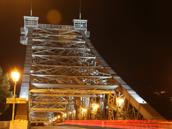 Die Loschwitzer Brücke – das "Blaue Wunder" – in Dresden, fotografiert bei Nacht, wird durch zahlreiche Strahler illuminiert. Die Laternen für die Fußwege folgen dem Straßenverlauf ebenso wie die roten Lichtspuren eines über die Brücke fahrenden Fahrzeuges.