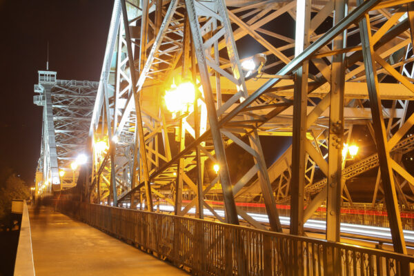 Die Loschwitzer Brücke – das "Blaue Wunder" – in Dresden, fotografiert bei Nacht, wird durch zahlreiche Strahler illuminiert. Der Blick folgt dem Geländer der stromabwärts liegenden Fußwegkonstruktion einerseits und den Laternen zur Fußwegbeleuchtung andererseits. Über den Fußweg spazieren Personen, die durch die Langzeitbelichtung jedoch nur schemenhaft erkennbar sind. Durch die sich rechts daneben erhebende Fachwerkskonstruktion der Brücke sind weiße und rote Lichtspuren über die Brücke fahrender Fahrzeuge zu erkennen.