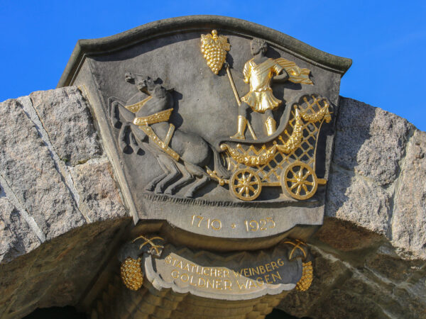 Das Wappen über dem Tor zum staatlichen Weinberg „Goldener Wagen“ glänzt in wunderbarem Gold und reflektiert das Sonnenlicht, zeigt aber auch einen angenehmem Kontrast zum matten Sandstein des Wappens. Darüber strahlt der Himmel tiefblau. Das Wappen zeigt einen Wagen, gezogen von zwei Pferden, auf dem ein Mann mit einem Stab posiert, an dem eine große Weintraube hängt.