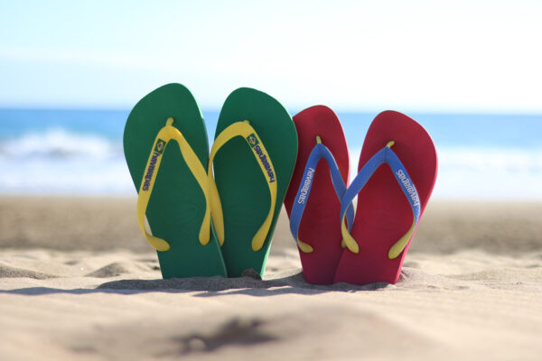 Grün-gelbe und rot-blaue Flip-Flops stecken im Sand am Playa de Maspalomas auf Gran Canaria. Nur unscharf ist dahinter das Wasser des Atlantik und der Himmel zu sehen.