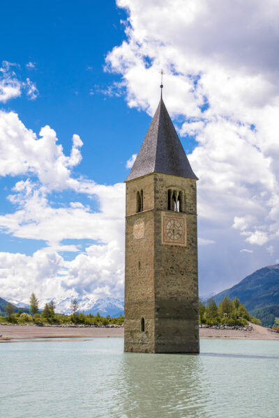 Der Kirchturm einer bei der Errichtung des Stausees aufgegebenen Siedlung ragt aus der Wasseroberfläche des Reschensees am Ende des Etschtals in Südtirol hervor.