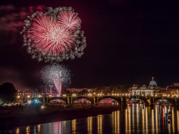 Hinter der elbüberspannenden Augustusbrücke steigt das Abschlussfeuerwerk zum "Canaletto"-Stadtfest in den Dresdner Nachhimmel. Über einer roten Feuerwerksfontäne auf der linken Bildseite explodieren mehrere Feuerwerkskörper und malen mit Hilfe einer längeren Belichtungszeit ebenfalls rote Lichtspuren in den Himmel. Am Rande der roten Lichtspuren der Fontäne und der am Himmel explodierenden Feuerwerkskörper folgt ein kleiner, weißer Funkenregen. Auf der rechten Bildseite ist die Kunstakademie zu sehen und die Lichter der Brühl'schen Terrasse und der Augustusbrücke reflektieren sich in der Elbe.