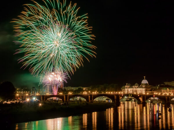 Hinter der elbüberspannenden Augustusbrücke steigt das Abschlussfeuerwerk zum "Canaletto"-Stadtfest in den Dresdner Nachhimmel. Über einer orange-lila-farbenen Feuerwerksfontäne auf der linken Bildseite explodieren mehrere Feuerwerkskörper und malen mit Hilfe einer längeren Belichtungszeit grün-orange und grün-pinke Lichtspuren in den Himmel. Auf der rechten Bildseite ist die Kunstakademie zu sehen und die Lichter der Brühl'schen Terrasse und der Augustusbrücke reflektieren sich in der Elbe.