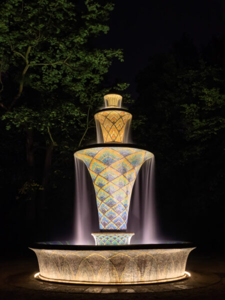 Der aus drei Wasserebenen bestehende Mosaikbrunnen im Großen Garten von Dresden ist märchenhaft beleuchtet. Das Wasser fließt von der oberen Ebene über die zwei weiteren bis in das Wasserauffangbecken. Durch die Langzeitbelichtung ist die Dynamik des Wassers gut eingefangen. Die mit Mosaiken in Schuppen angeordneten Farbverläufe von braun-orange-gelb, über blau-türkis-orange bis hin zu lila-blau-türkis strahlen wunderbar farbkräftig. Um den Brunnen herum ist es tiefste Nacht. Nur ein paar wenige belaubte Baumzweige sind im Hintergrund erkennbar.