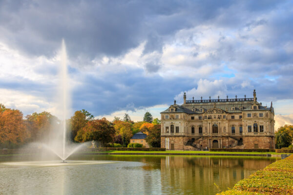 Es ist Herbst und wechselhaftes Wetter. Die Wolken zeichnen eine sehr interessante Struktur mit gutem Kontrast und Farben von gelb bis dunklerem Blau in den Himmel. An einigen Stellen ist auch hellblauer Himmel zu sehen. Darunter liegt das Palaisschlösschen im Großen Garten von Dresden. Die Bäume rechts und links davon weisen abwechslungsreiche Herbstlaubfärbung auf. Im Vordergrund liegt der Palaisteich und der Springbrunnen darin schießt eine meterhohe Fontäne in die Luft.