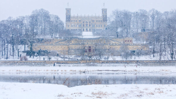 Das Schloss Albrechtsberg als eines der drei Elbschlösser Dresdens liegt am Hang des nordelbischen Ufers, fotografiert vom Elberadweg auf der altstädter Seite. Die Szenerie ist komplett verschneit, Schneeflocken tanzen wild durch das gesamte Bild. Auf dem Elberadweg unterhalb des Schlosses sind Skilangläufer und davor die Elbe zu sehen.