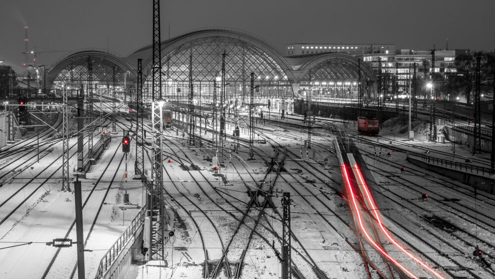 Der Blick fällt von der Budapester Brücke aus auf den Dresdner Hauptbahnhof. Ein einfahrender Zug hinterlässt zwei rote Lichtspuren. Rote Lichtquellen und zwei rote Lokomotiven sind im Bild erkennbar. Im Übrigen ist das Bild schwarz-weiß. Es hat geschneit und das Bahnhofsvorfeld mit seinem Schienennetz zeigt einen schönen Kontrast.