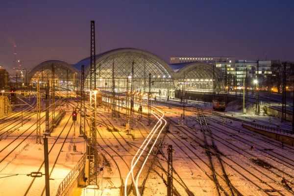 Der Blick fällt von der Budapester Brücke aus auf den Dresdner Hauptbahnhof in der blauen Stunde. Ein ausfahrender Zug hinterlässt drei gelbe Lichtspuren. Es hat geschneit und je nach reflektiertem Licht erstrahlt das Bahnhofsvorfeld mit seinem Schienennetz in den Farben gelb bis lila. Der Himmel hat eine lila Farbgebung.