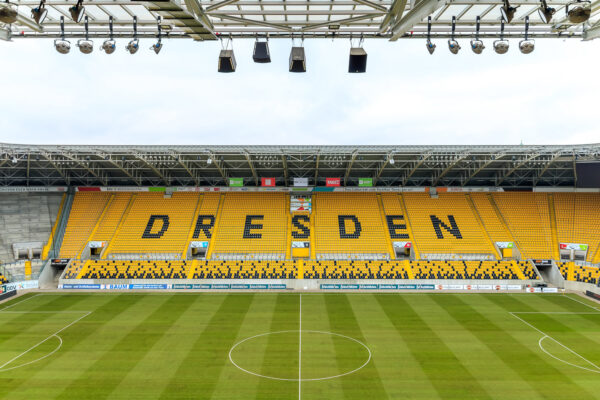 Von der Pressetribüne direkt unterm Stadiondach oberhalb des VIP-Bereiches des Rudolf-Harbig-Stadions in Dresden fällt der Blick auf die Gegengerade mit dem aus Sitzschalen geformten "DRESDEN"-Schriftzug. Am oberen Bildrand ist noch das Stadiondach von der VIP-Seite mit Lautsprechern und Flutlichtstrahlern zu sehen. Vor der Gegengerade liegt der sattgrüne Rasen des Fußballfeldes.