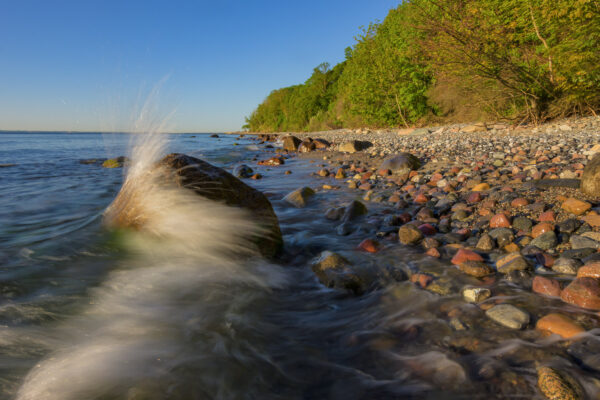 Die Sonne ist gerade aufgegangen. Am steinigen Strand von Kap Arkona auf Rügen bricht sich eine Welle an einem etwa halben Meter hohen Findling im Wasser.